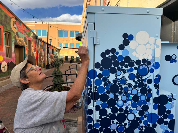 Artist painting mural on cabinet with colorful wall mural and building with windows in the background.