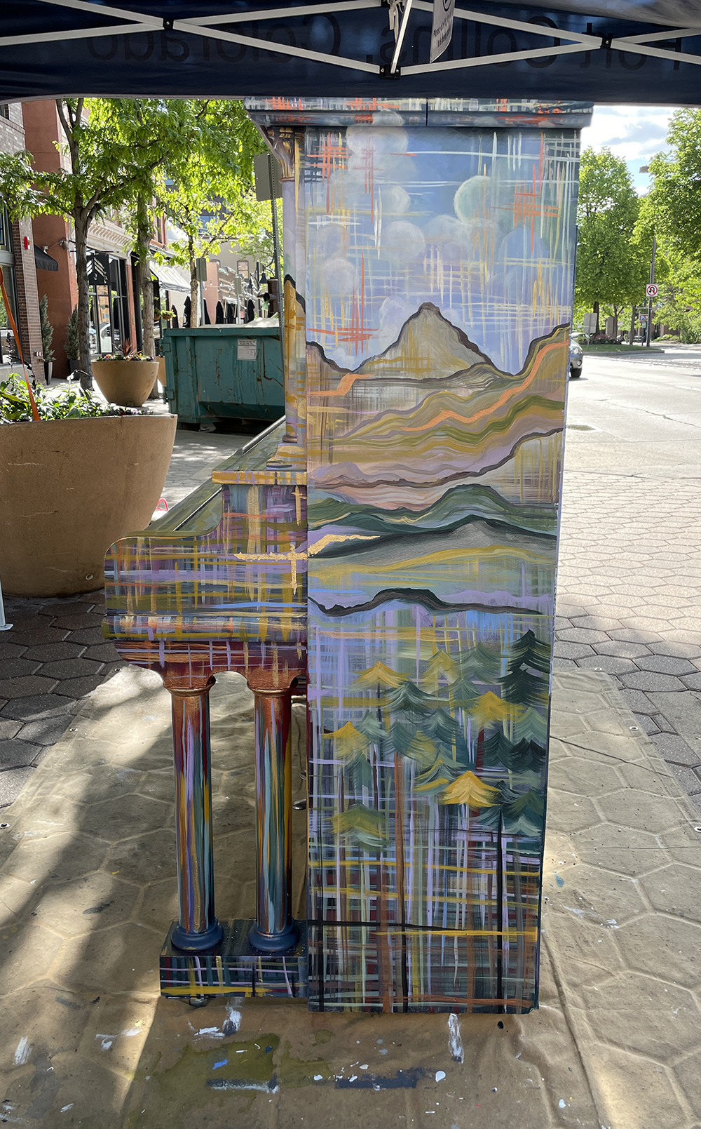 side of painted piano with flower pot and teal dumpster in the back.