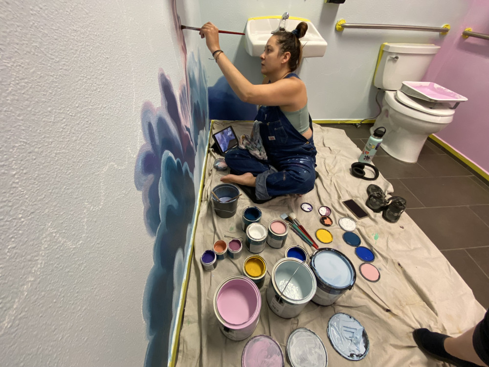 artist sitting on the floor painting a mural on the wall with a sink and toilet behind her