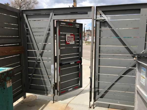Photo taken inside a trash enclosure, looking out a pedestrian access door. There are two dumpsters inside the enclosure.