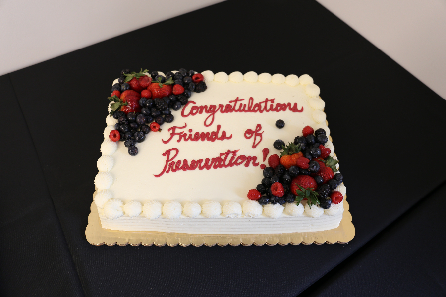 White cake on black table; cake reads "Congratulations Friends of Preservation"