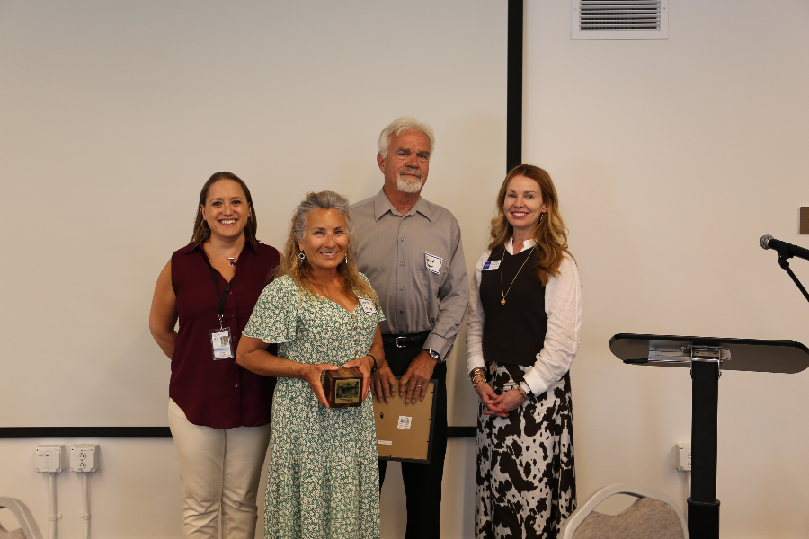 Four individuals stand in front of a screen; one holds a square wood award block