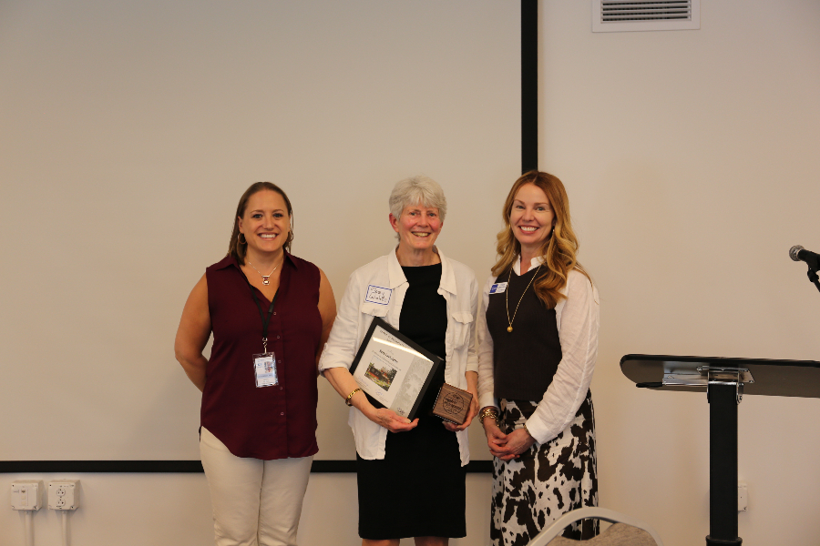 Three individuals stand in front of a blank screen; one holds a picture frame and a square wood award block