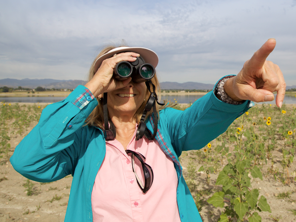 Woman on a warm, summer day smiling and pointing towards the camera as she peers through binoculars.