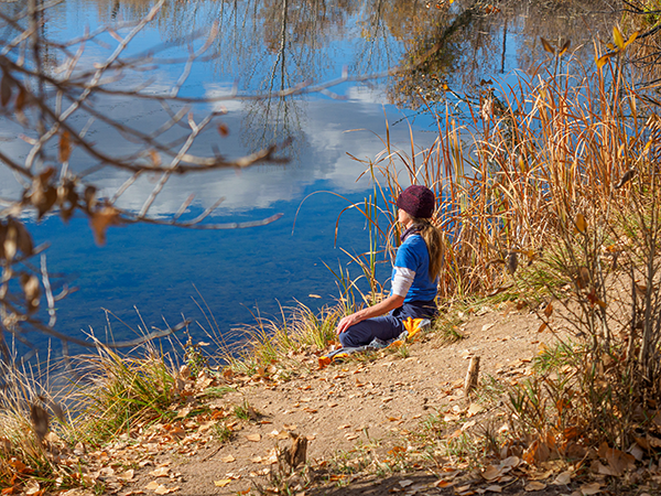 Woman meditating peacefully, sitting on the ground at the edge of a pond at McMurry Natural Area in Fort Collins.