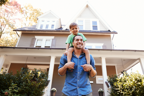 Photograph of a person standing outside of a house with a child on their shoulders. The adult and child are both smiling.