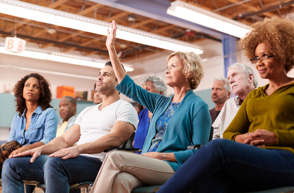 Group of adults in a room, sitting in chairs and looking in the same direction. A blonde woman has her hand raised.