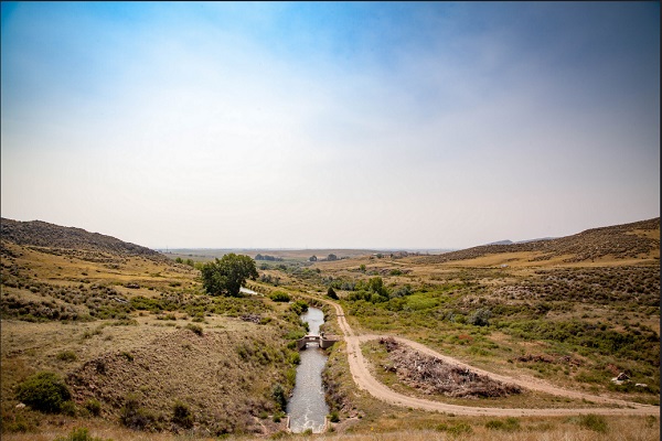 A river splits the middle of a dusty valley with a dirt road and large green bush beside it. A small bridge in the middle.